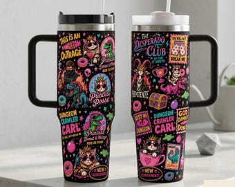May include: Two black stainless steel tumblers with handles and straws, featuring colorful graphic prints of cats, dinosaurs, donuts, and text including "This is an Outrage," "Princess Posse," "The Desperado Club," and "Dungeon Crawler Carl."