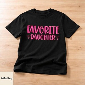 May include: Black t-shirt with the text "FAVORITE DAUGHTER" in pink, with bow accents. The shirt is made of a soft material and is suitable for casual wear. The design is centered on the front of the shirt.
