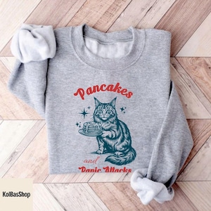 May include: A gray sweatshirt with the words "Pancakes" and "Panic Attacks" in red script. A detailed illustration of a cat holding a plate of pancakes is featured below the word "Pancakes". The sweatshirt has long sleeves.