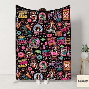 May include: A black fleece blanket featuring a repeating pattern of Dungeon Crawler Carl characters, including cats wearing crowns, a goat, and a dinosaur. The design includes colourful donuts, skulls, bombs, and text like The Desperado Club.