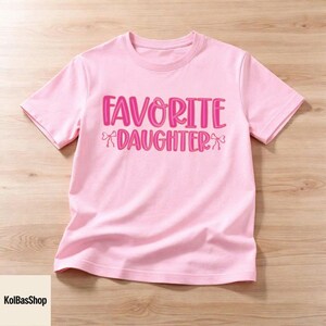 May include: A light pink t-shirt with the words "FAVORITE DAUGHTER" in bright pink, with bow accents. The shirt is made of soft material and is displayed on a light wood surface. The text is in a playful, bold font.