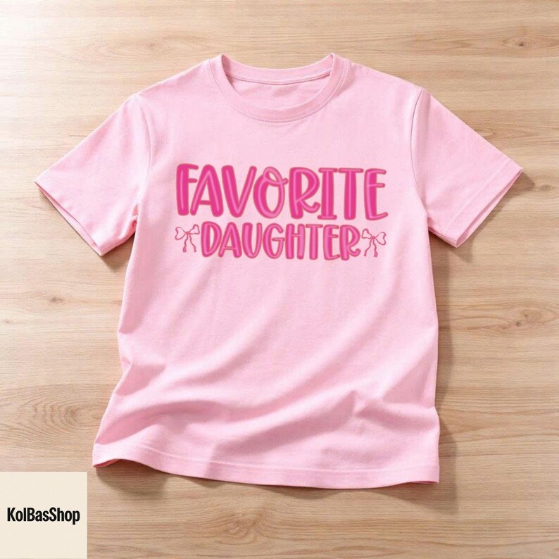 May include: A light pink t-shirt with the words "FAVORITE DAUGHTER" in bright pink, with bow accents. The shirt is made of soft material and is displayed on a light wood surface. The text is in a playful, bold font.