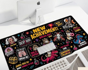 May include: A large black rectangular desk mat featuring a colorful graphic print with the text NEW ACHIEVEMENT, DESPRADO, PRINCESS POSSE, YOO HOO, HIII ZEV, VIVA LA REVOLUTION CARL, MONGO IS APPALLED, I AM YELLING CARL, and DUNGEON CRAWLER alongside various illustrations.