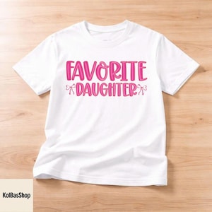 May include: White t-shirt with the text "FAVORITE DAUGHTER" in pink, with bow accents. The shirt is made of a soft material and is suitable for casual wear. The design is centered on the front of the shirt.