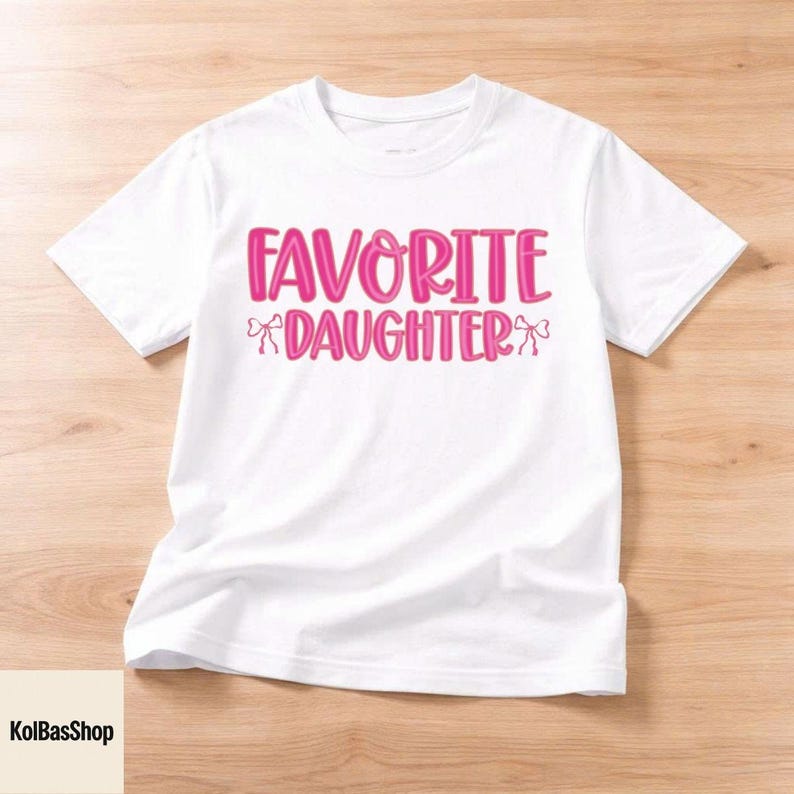 May include: White t-shirt with the text "FAVORITE DAUGHTER" in pink, with bow accents. The shirt is made of a soft material and is suitable for casual wear. The design is centered on the front of the shirt.