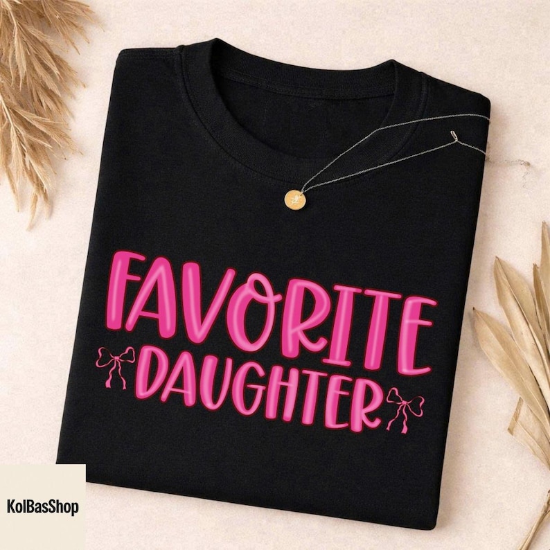 May include: Black t-shirt with the text "FAVORITE DAUGHTER" in pink, with bow accents. A gold pendant necklace rests on the shirt. The KolBasShop logo is in the bottom left corner.