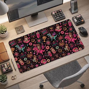 May include: A rectangular desk mat featuring a black background with a repeating pattern of pink dinosaurs, cats wearing crowns, donuts, and beverage cups. The design includes text such as Princess Donut Mongo, Ride or Roar, and Damnit Donut.
