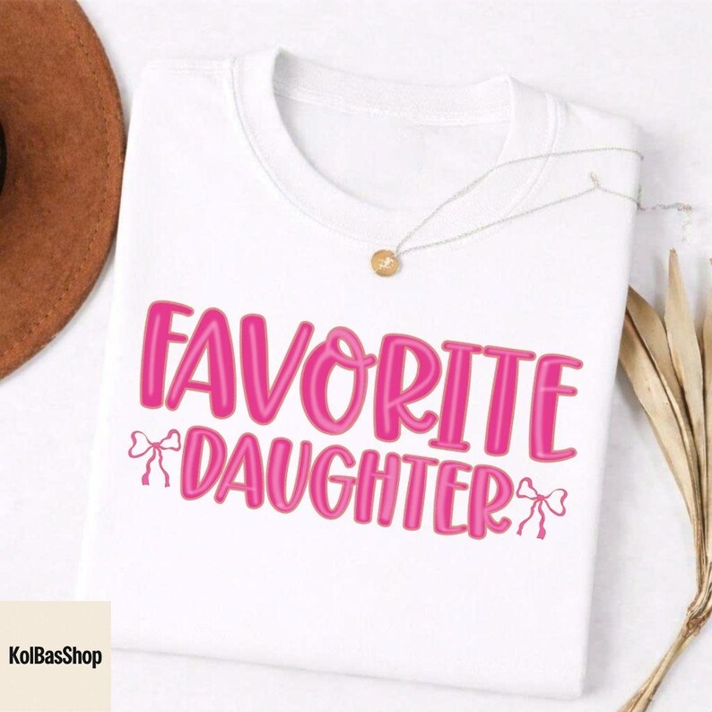May include: White t-shirt with the text "FAVORITE DAUGHTER" in bright pink, accented with bow graphics. A gold pendant necklace and a brown hat are visible in the background. The shirt is a casual wear item.