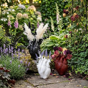 May include: Three heart-shaped vases, black, white, and burgundy, each filled with different dried flowers and foliage. The vases are placed on a stone path in a garden setting, surrounded by greenery and flowers.