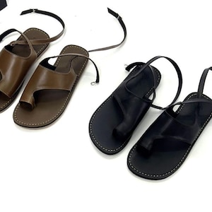 Op de afbeelding: Twee paar leren sandalen. Eén paar is bruin, het andere is zwart. Elke sandaal heeft een teengreep en een enkelband. De sandalen staan op een witte achtergrond.