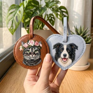 Può includere: Un'etichetta per bagagli rotonda in pelle marrone con un gatto tigrato dipinto che indossa una corona di fiori rosa e un'etichetta a forma di cuore in pelle azzurra con un cane border collie bianco e nero dipinto dall'espressione felice.