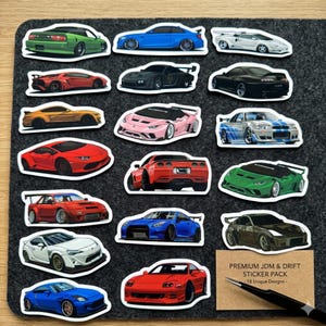 Puede incluir: Un conjunto de 18 pegatinas únicas de coches JDM y drift en varios colores, incluyendo verde, azul, rojo y negro. Las pegatinas están dispuestas sobre una superficie gris oscuro. El paquete de pegatinas incluye una variedad de modelos de coches.