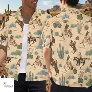 May include: A short-sleeved button-down shirt with a beige background featuring a repeating pattern of cowboys on horseback, cacti, and desert plants. The shirt is worn open over a white undershirt. The clothing shop logo is visible in the bottom left corner.