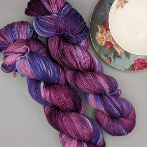 Op de afbeelding: Twee strengen handgeverfd garen in tinten paars, roze en blauw. Het garen ligt naast een vintage kop en schotel met bloemmotieven. Het garen ziet er zacht uit en is klaar voor brei- of haakprojecten.