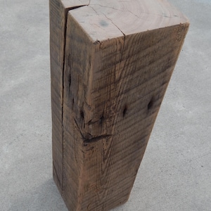 May include: A tall, rectangular wooden block with a weathered brown finish. The top surface displays visible circular growth rings and a deep vertical crack extending down one side. The wood grain is textured with natural knots and fissures.