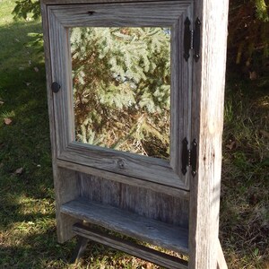 May include: A rustic wooden cabinet with a mirror door and a shelf. The cabinet is made of reclaimed wood and has a gray finish. It has black metal hinges and a knob.