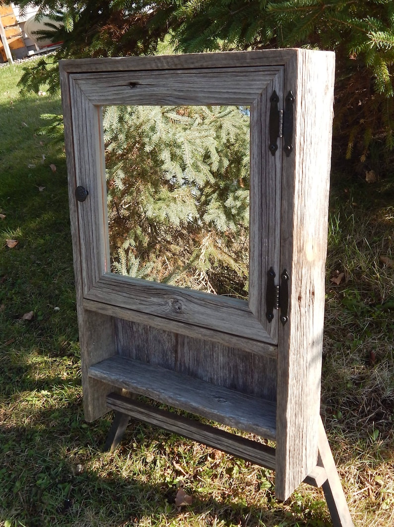 May include: A rustic wooden cabinet with a mirror door and a shelf. The cabinet is made of reclaimed wood and has a gray finish. It has black metal hinges and a knob.