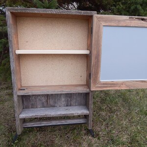 May include: A rustic wooden cabinet with a hinged door and shelves. The cabinet has a light gray painted interior and a distressed wood finish. The cabinet is standing on black metal legs.