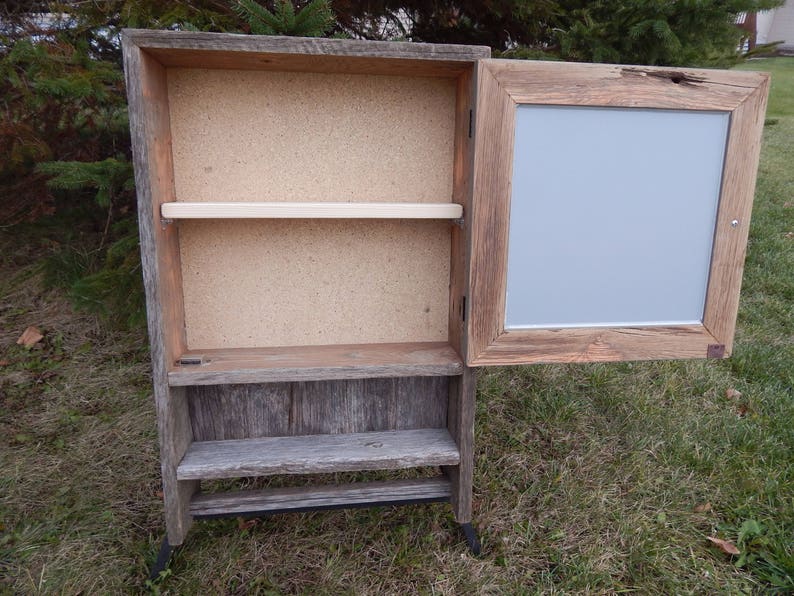 May include: A rustic wooden cabinet with a hinged door and shelves. The cabinet has a light gray painted interior and a distressed wood finish. The cabinet is standing on black metal legs.