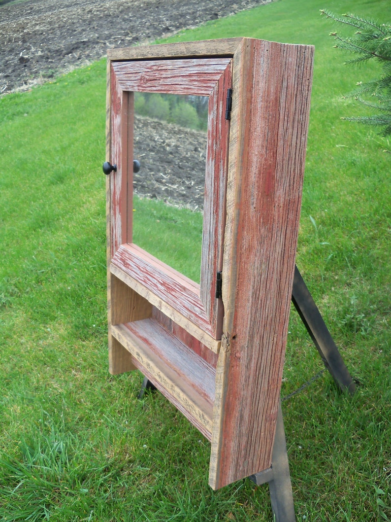 May include: A rustic wooden medicine cabinet with a red stained finish and a mirror door. The cabinet has a small shelf inside and a black knob.