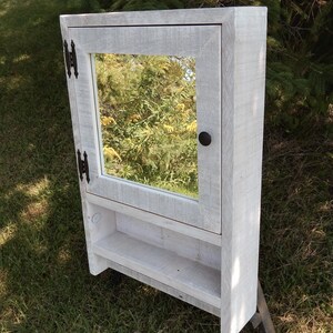 May include: White wooden bathroom cabinet with a mirror and a shelf. The cabinet has black hinges and a black knob.