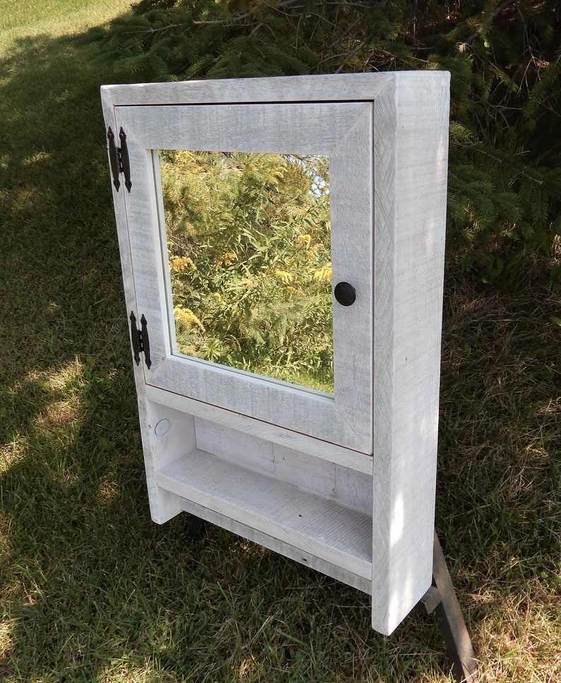 May include: White wooden bathroom cabinet with a mirror and a shelf. The cabinet has black hinges and a black knob.
