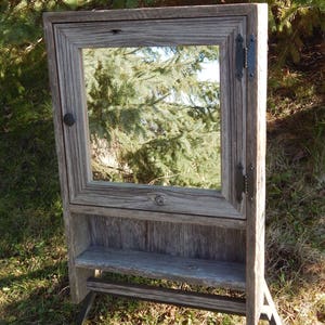 May include: A rustic wooden bathroom cabinet with a mirror, a shelf, and a towel bar. The cabinet is made of reclaimed wood and has a weathered gray finish. The mirror is framed in wood and has a simple design. The shelf is made of wood and is perfect for storing toiletries. The towel bar is made of metal and is attached to the side of the cabinet.