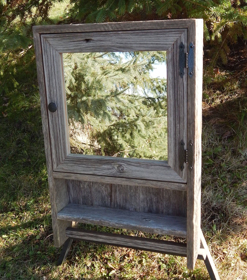 May include: A rustic wooden bathroom cabinet with a mirror, a shelf, and a towel bar. The cabinet is made of reclaimed wood and has a weathered gray finish. The mirror is framed in wood and has a simple design. The shelf is made of wood and is perfect for storing toiletries. The towel bar is made of metal and is attached to the side of the cabinet.