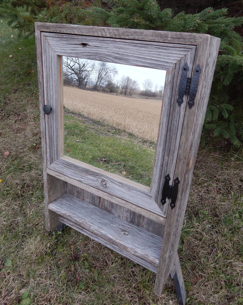 May include: A rustic wooden mirror with a shelf, featuring a gray weathered finish and black metal hinges. The mirror reflects a view of a field.
