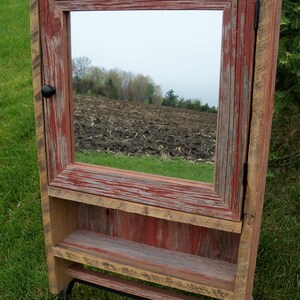 May include: Rustic wooden bathroom cabinet with a mirror, a shelf, and a towel bar. The cabinet is painted in shades of red and brown, with a distressed finish. The mirror reflects a view of a field.
