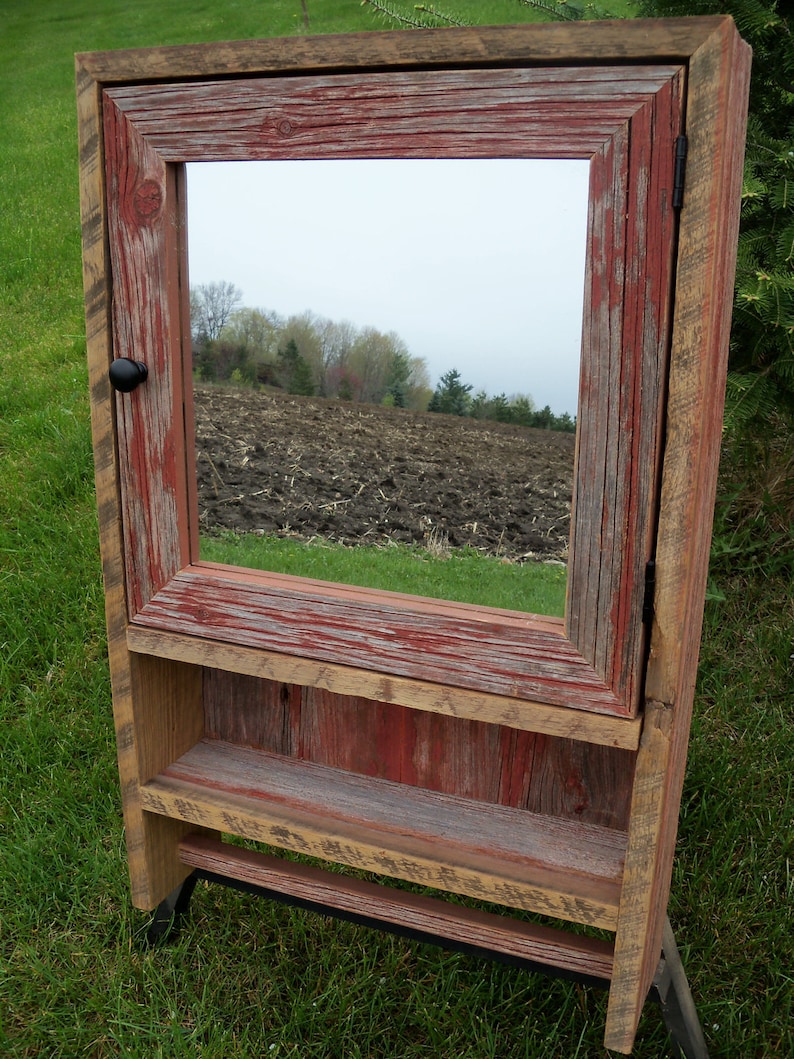 May include: Rustic wooden bathroom cabinet with a mirror, a shelf, and a towel bar. The cabinet is painted in shades of red and brown, with a distressed finish. The mirror reflects a view of a field.