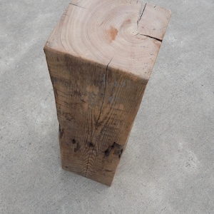 May include: A tall, square-cut wooden pillar with a visible grain pattern on the top surface. The wood features a light brown, weathered texture with natural cracks and knots running vertically along its sides, showing a raw, unfinished appearance.