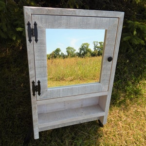 May include: White wooden bathroom cabinet with a mirror and a shelf. The cabinet has black metal hinges and a black knob.