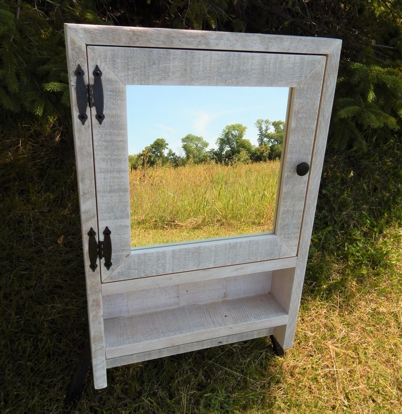 May include: White wooden bathroom cabinet with a mirror and a shelf. The cabinet has black metal hinges and a black knob.
