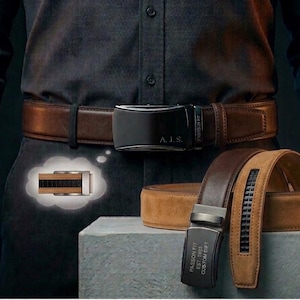 Op de afbeelding: Een bruine leren riem met een zwarte gesp met de initialen A.J.S. en de tekst PASSION FIT EST. 1888 CUSTOM DRIFT. Een tweede beige suède riem met een zwarte gesp en een zwarte centrale strook ligt opgerold op een grijs blok.