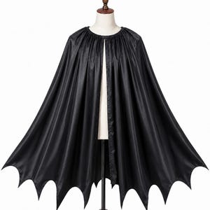 May include: A black cape featuring a gathered neckline and a hemline cut into distinct, pointed bat-wing shapes. The garment is made of a smooth, slightly reflective fabric and hangs open at the front, displaying a full, flowing silhouette.