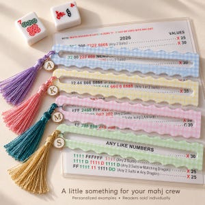 May include: Four scalloped-edge plastic Mah Jongg card holders in light blue, yellow, pink, and green gingham patterns. Each features a matching colored tassel and a gold-tone charm with a letter. They are placed over a 2026 Mah Jongg score card.