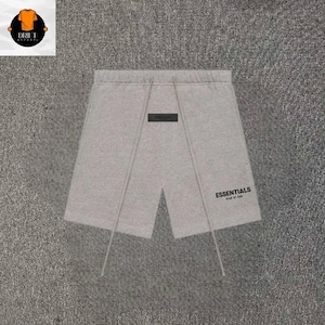 Peut inclure: Short gris clair doté d'une taille élastique avec de longs cordons de serrage pendants. Un petit écusson rectangulaire noir est centré sous la taille, et le mot ESSENTIALS est imprimé en noir sur la jambe droite du vêtement.