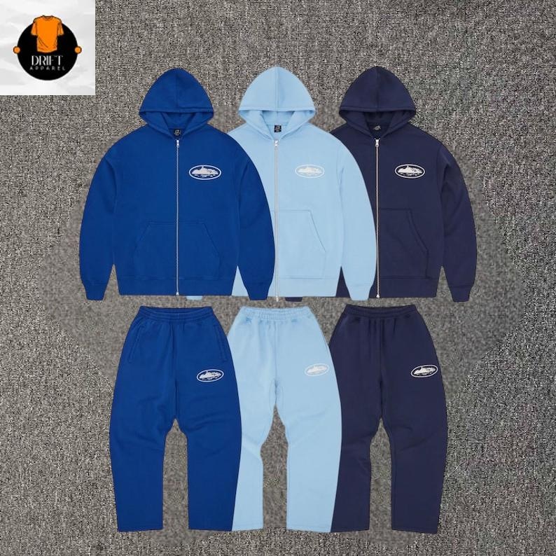 Puede incluir: Tres conjuntos a juego de sudaderas con cremallera y pantalones de ch&aacute;ndal en azul real, azul claro y azul marino. Cada prenda presenta un peque&ntilde;o gr&aacute;fico ovalado blanco en el pecho y el muslo con un dibujo de un coche y el texto Drift Apparel.