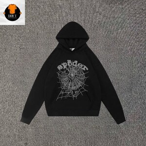 May include: A black long-sleeve hooded sweatshirt featuring a large, light gray graphic print on the front. The graphic depicts a detailed spider web design with the word "sp&ouml;der" written in a stylized, rounded font arched above the web.