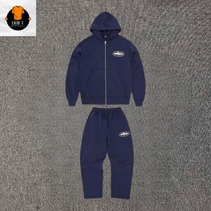 Puede incluir: Un conjunto de sudadera con capucha y cremallera y pantalones de ch&aacute;ndal en color azul marino oscuro. La sudadera tiene una cremallera plateada y un gr&aacute;fico ovalado blanco en el pecho izquierdo. El pantal&oacute;n incluye un gr&aacute;fico ovalado a juego.