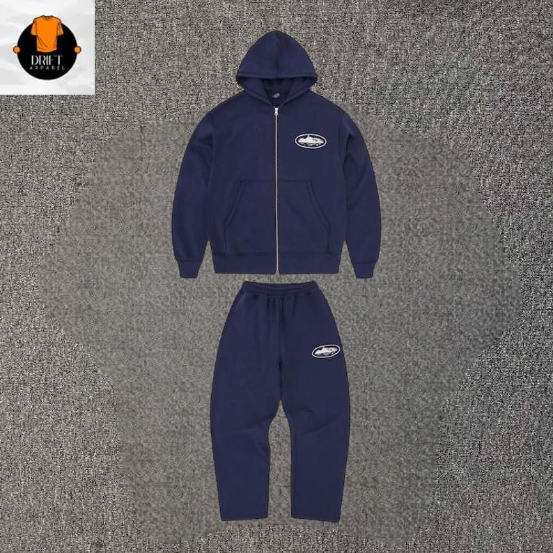 Puede incluir: Un conjunto de sudadera con capucha y cremallera y pantalones de ch&aacute;ndal en color azul marino oscuro. La sudadera tiene una cremallera plateada y un gr&aacute;fico ovalado blanco en el pecho izquierdo. El pantal&oacute;n incluye un gr&aacute;fico ovalado a juego.