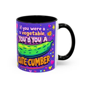 May include: White ceramic mug with a black handle and interior. The mug features a purple background with a green cucumber illustration and the text "If you were a vegetable, you'd you a Cute-Cumber."