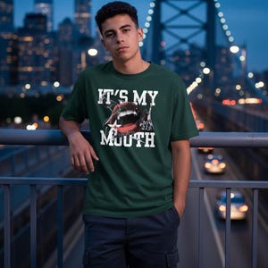 Può includere: T-shirt verde scuro con la scritta bianca "IT'S MY MOUTH" e una grafica di una bocca. La persona indossa la maglietta su un ponte con lo skyline della città sullo sfondo.