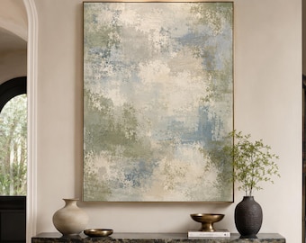 May include: A large rectangular abstract painting featuring blended shades of sage green, soft blue, and cream white, displayed in a thin gold frame. The artwork is mounted above a dark, textured stone console table with two thick pedestal legs.