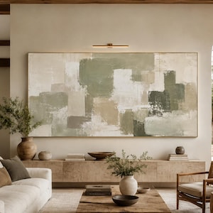 May include: A large rectangular abstract painting featuring a palette of sage green, beige, cream, and dark olive tones with visible brushstrokes. The artwork is displayed horizontally on a wall above a long wooden console table in a living room.