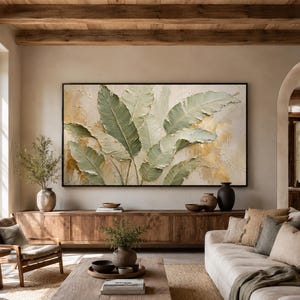 May include: A large rectangular wall art piece featuring a textured impasto painting of sage green banana leaves against a cream and gold background, framed in a thin black border, displayed horizontally above a wooden console table.