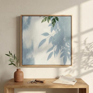May include: Square framed wall art featuring a light blue and white abstract design with leaf shadows. The artwork is in a natural wood frame. A small terracotta vase with greenery, keys, and a letter are on a wooden table.
