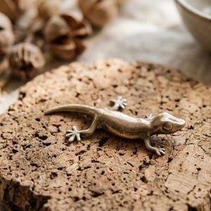 May include: A detailed, metallic gold-coloured lizard figurine with outstretched legs and a curved tail rests on a textured, brown cork surface. The lizard's features are clearly defined, showcasing its eyes and tiny feet.