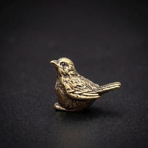 Może przedstawiać: Mała, mosiężna figurka ptaka. Ptak przedstawiony jest w pozycji siedzącej, z głową lekko uniesioną. Figurka ma teksturowane detale na skrzydłach i ciele, na ciemnym tle.